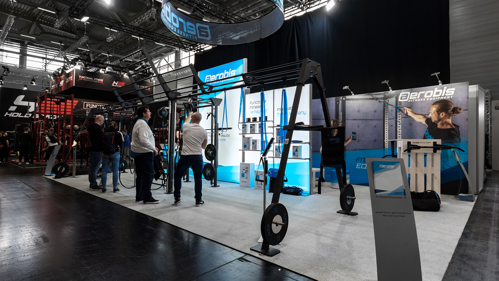 Auf einem Messestand werden Fitnessgeräte ausgestellt, die von mehreren Personen beobachtet werden, die miteinander kommunizieren.