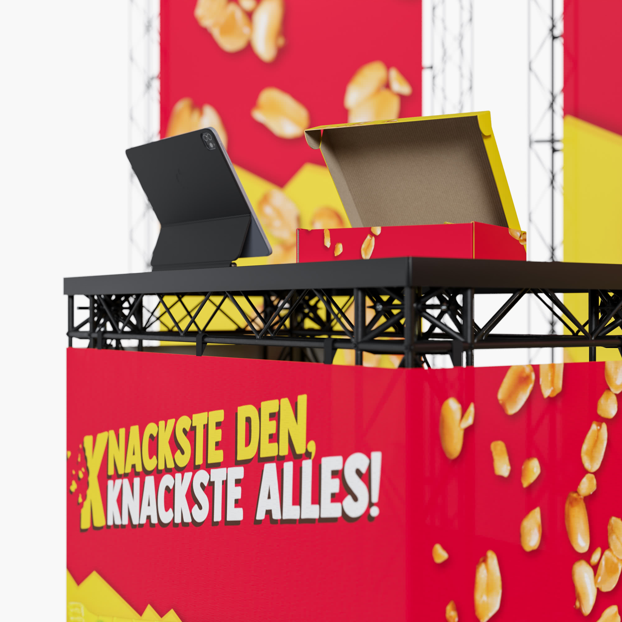Ein rot-gelbes Werbedisplay mit Erdnüssen und dem deutschen Text "Knackste den, knackste alles!" steht auf einem Tisch mit schwarzem Rahmen. Eine offene Pizzaschachtel und ein schwarzes elektronisches Gerät liegen darauf.