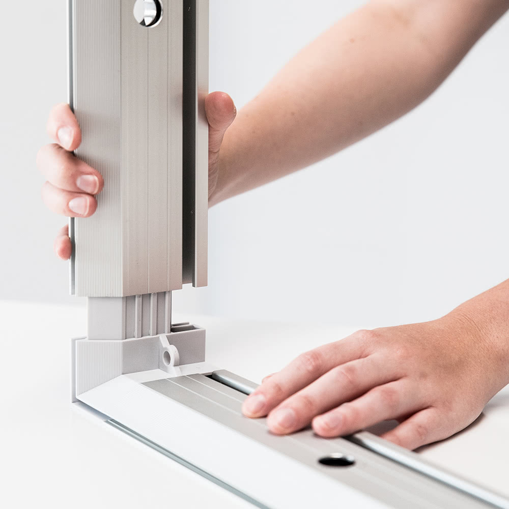 Eine Person baut einen Metallrahmen zusammen, indem sie ein vertikales Aluminiumteil in eine Eckverbindung auf einer weißen Fläche schiebt und dabei beide Hände zur Ausrichtung und Stabilität benutzt.
