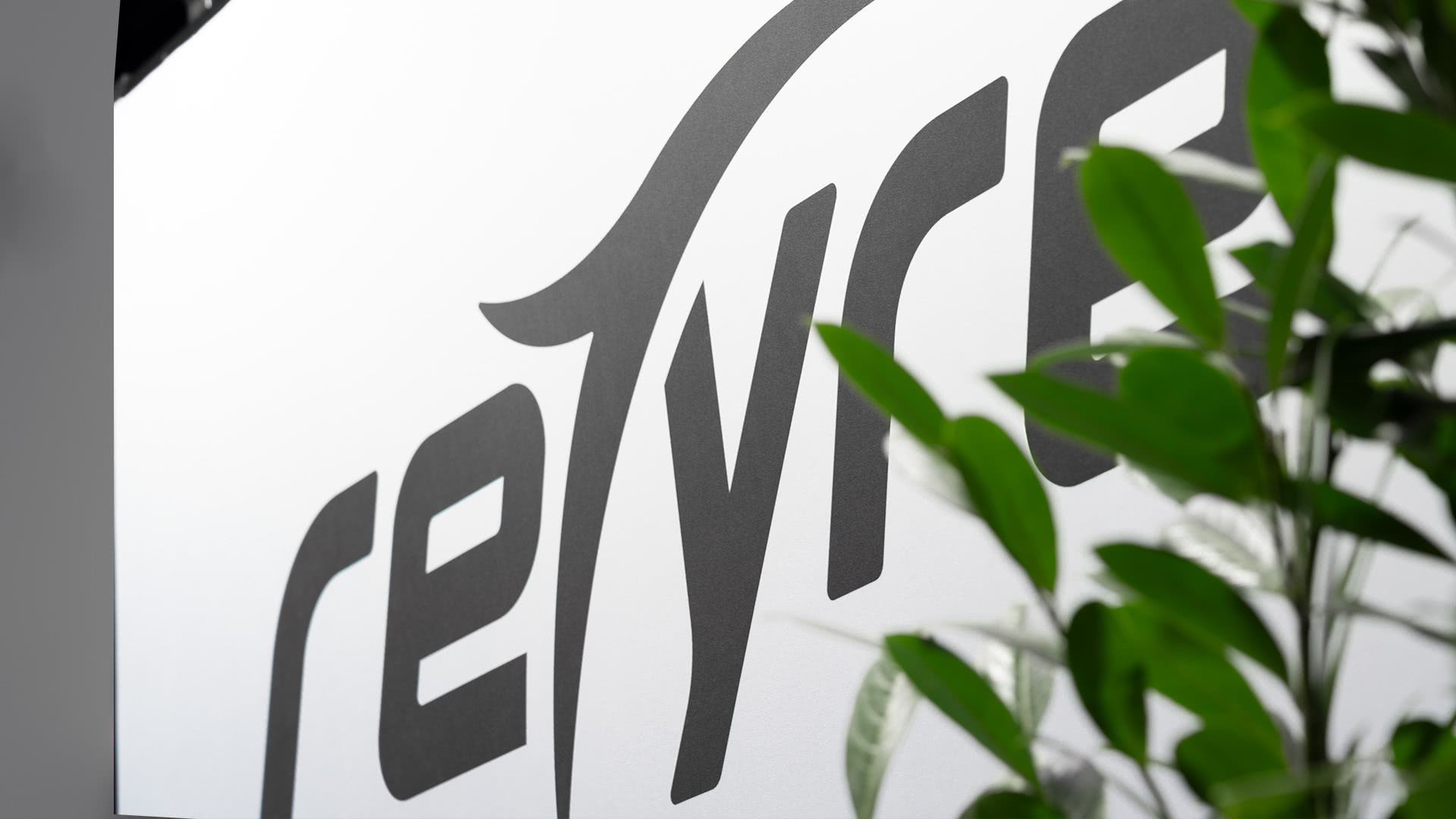 Nahaufnahme eines Wandschilds mit dem Wort "retyre" in fetten, stilisierten schwarzen Buchstaben, teilweise verdeckt durch grüne Blattpflanzen im Vordergrund.