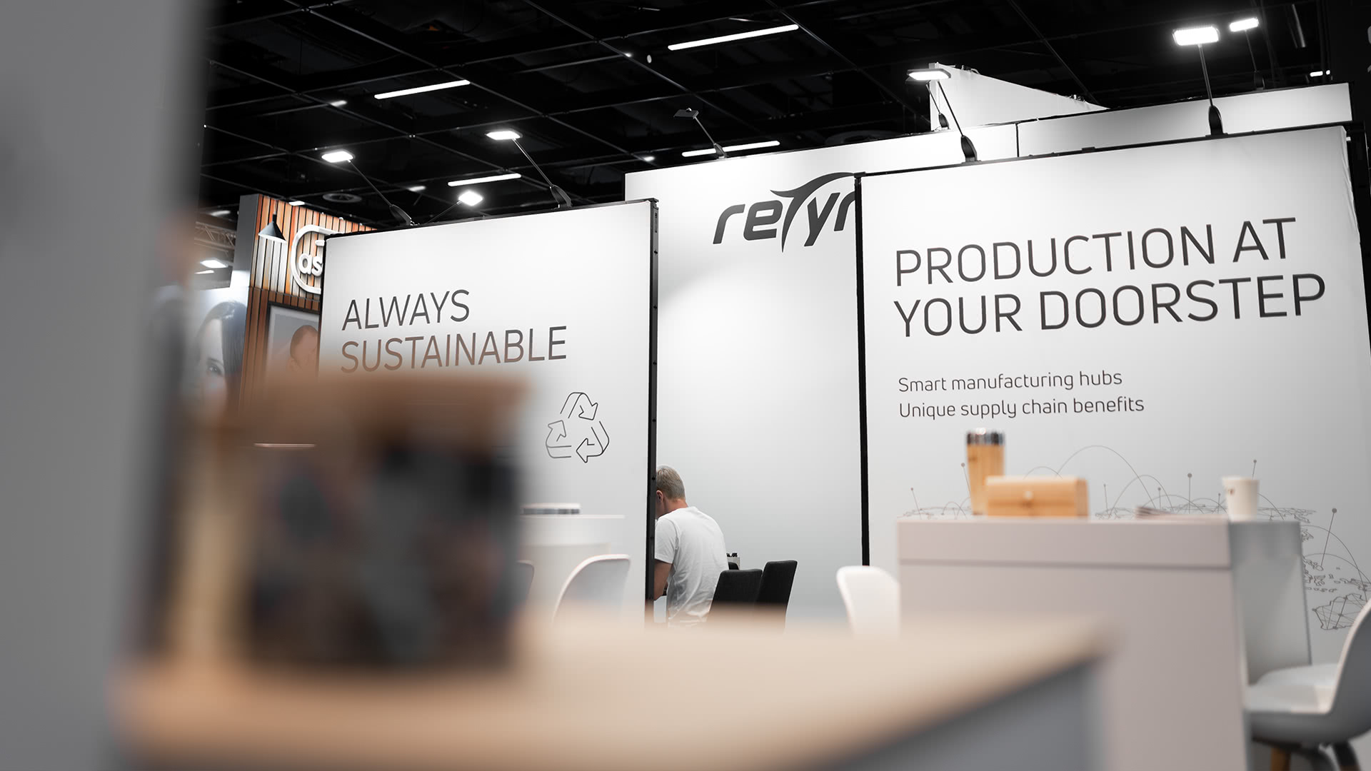 Ein Messestand mit weißen Wänden zeigt die Slogans ALWAYS SUSTAINABLE und PRODUCTION AT YOUR DOORSTEP. Eine sitzende Person ist teilweise sichtbar; der Vordergrund ist unscharf.