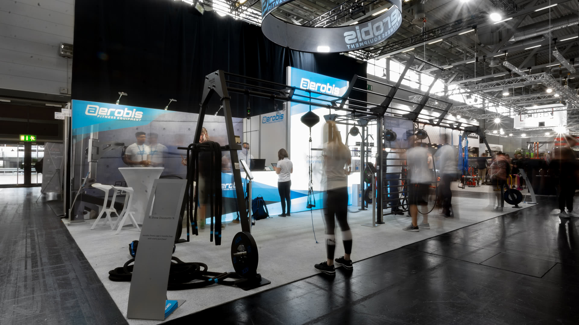 Messestand mit Fitnessgeräten und "Aerobis"-Branding, mit Menschen, die in einem hell erleuchteten Kongresszentrum interagieren, trainieren und herumlaufen. Einige Figuren erscheinen aufgrund der Bewegung unscharf.