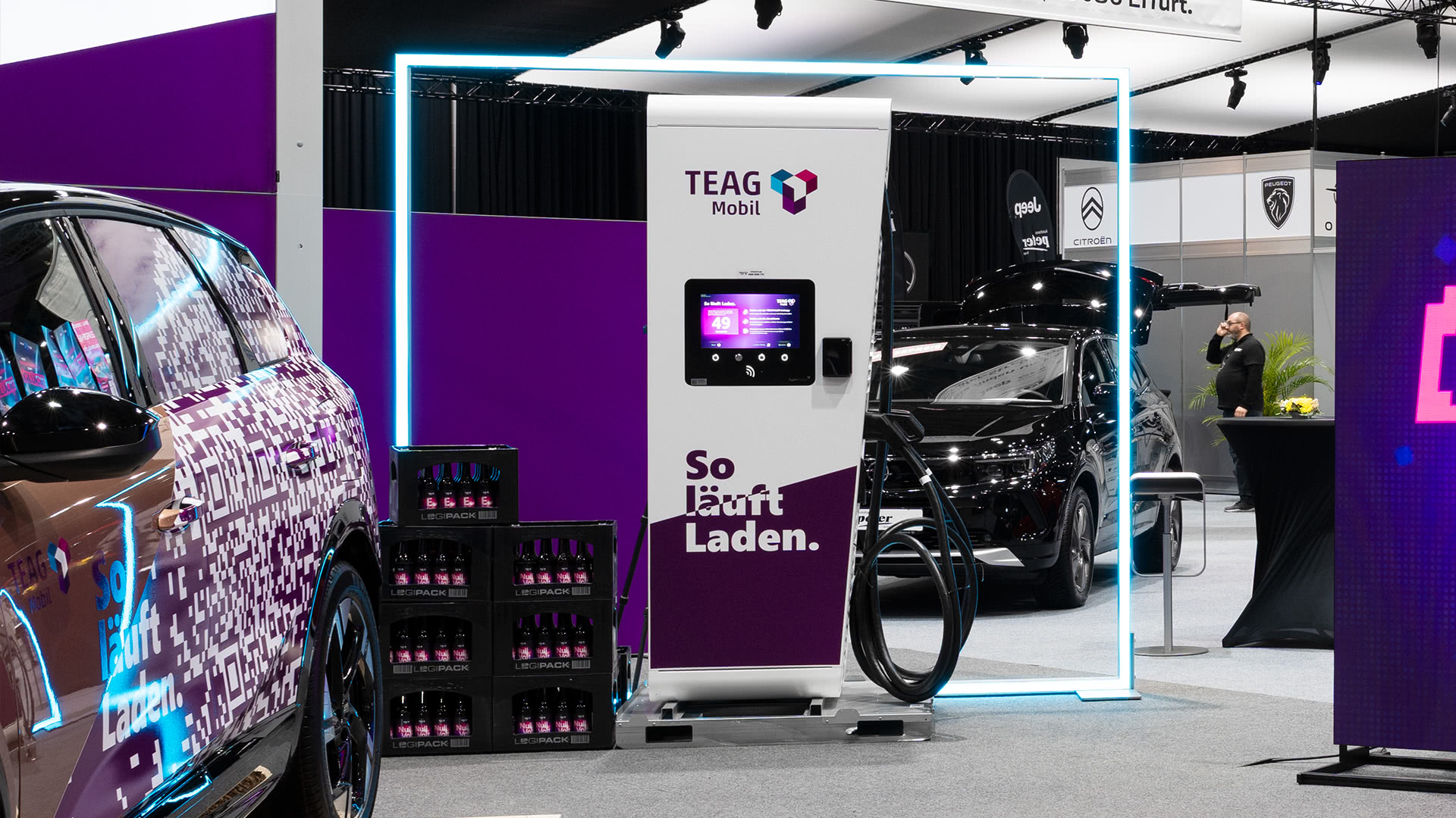 Eine moderne Ladestation für Elektrofahrzeuge der Marke TEAG Mobil steht in einer Messehalle, umgeben von geparkten Autos und lila beleuchteten Displays. Der Bildschirm des Ladegeräts ist sichtbar, zusammen mit mehreren Ladekabeln.