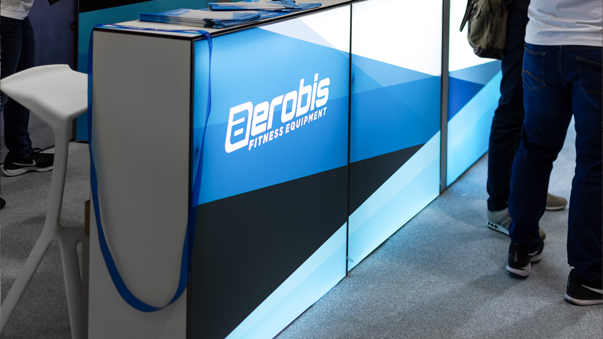 Eine moderne Ausstellungstheke mit dem Logo von Aerobis Fitness Equipment, dekoriert mit blauen, schwarzen und weißen geometrischen Mustern, mit Broschüren obenauf und Menschen, die daneben stehen.