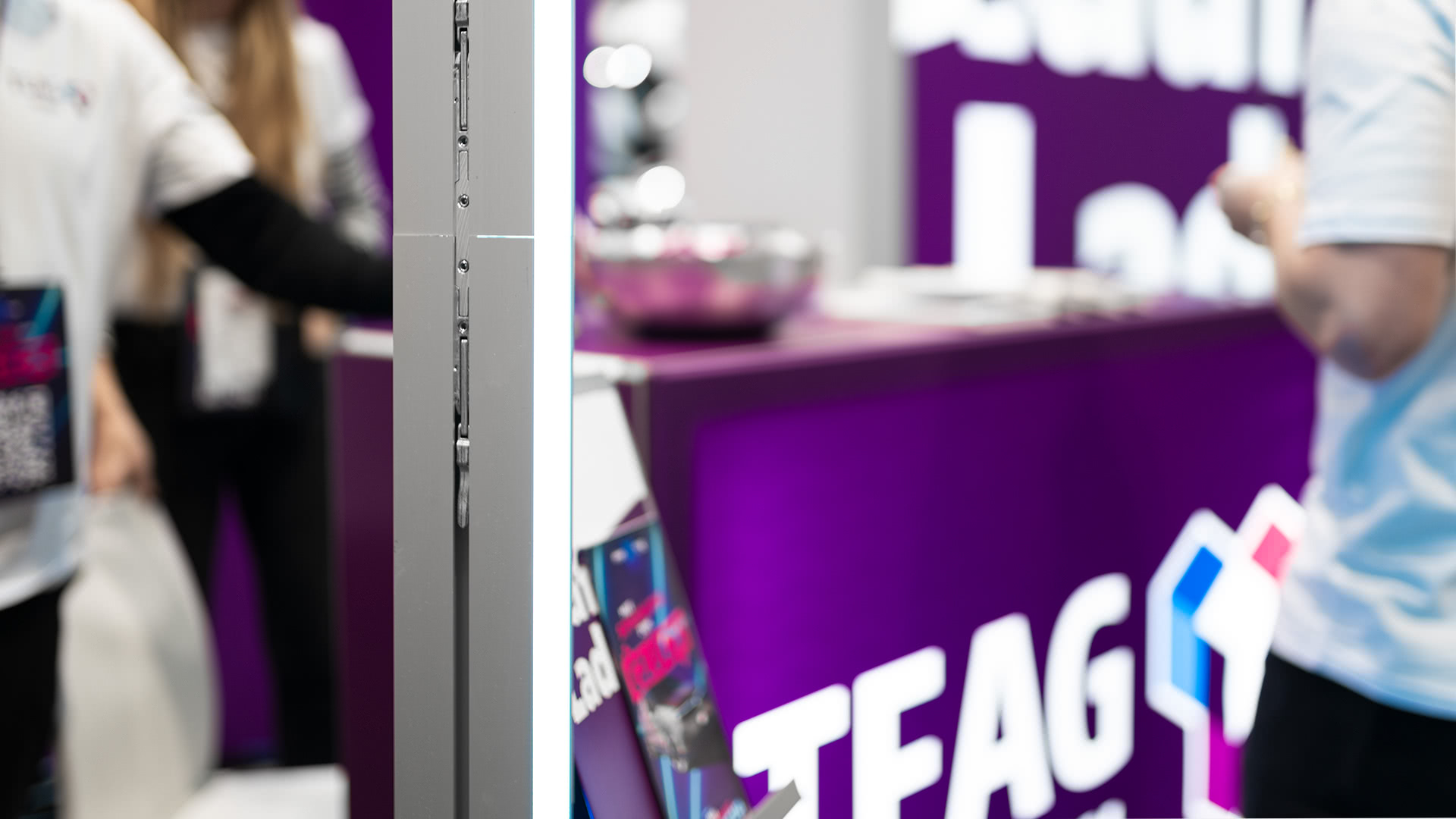 Im Hintergrund steht ein lilafarbener Messestand mit unscharfen Personen in weißen Hemden, während im Vordergrund eine Metalltrennwand und ein Prospektständer zu sehen sind. Auf der Theke ist ein Teil eines großen TEAG-Logos zu sehen.