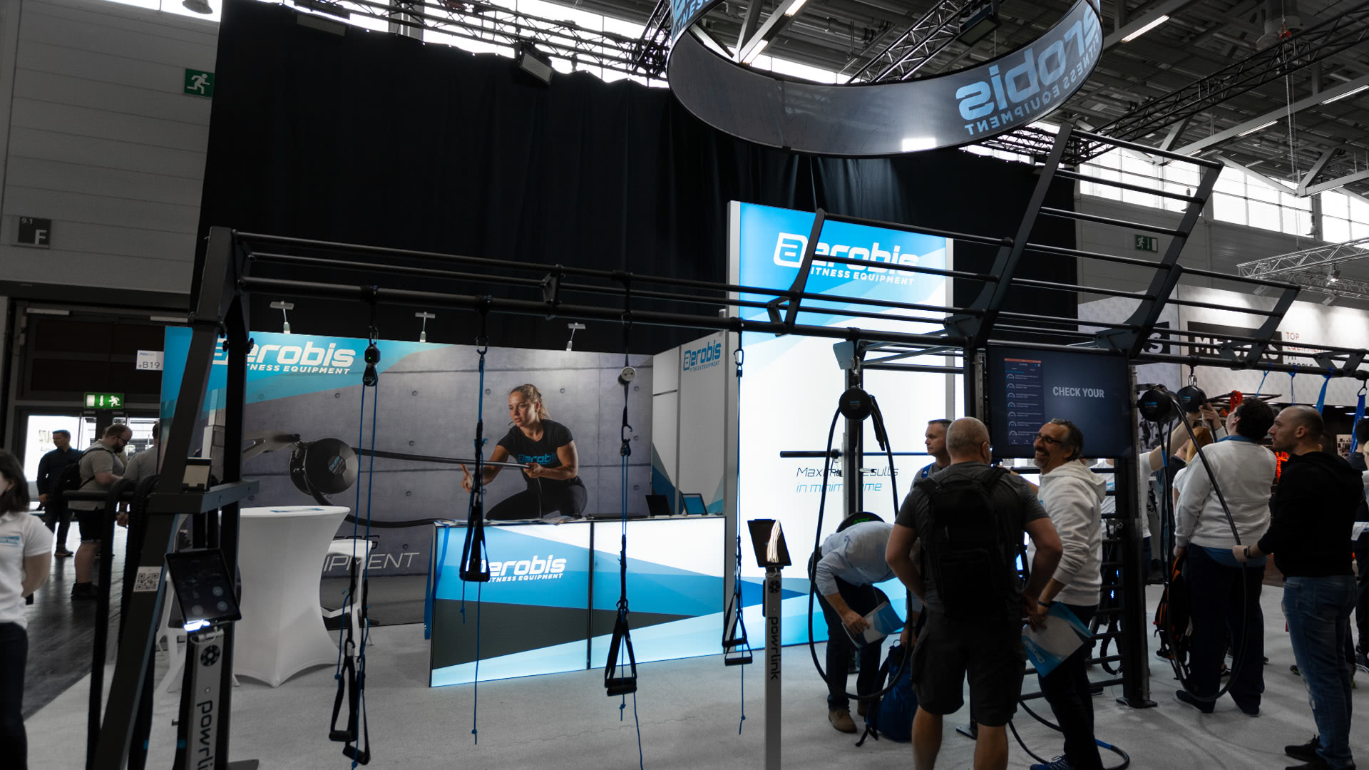 Ein Messestand für Aerobis zeigt Fitnessgeräte, Banner und einen Bildschirm mit einer Frau, die ein Trainingsgerät benutzt. Mehrere Personen beobachten die Ausstellung in einer großen Kongresshalle oder interagieren mit ihr.