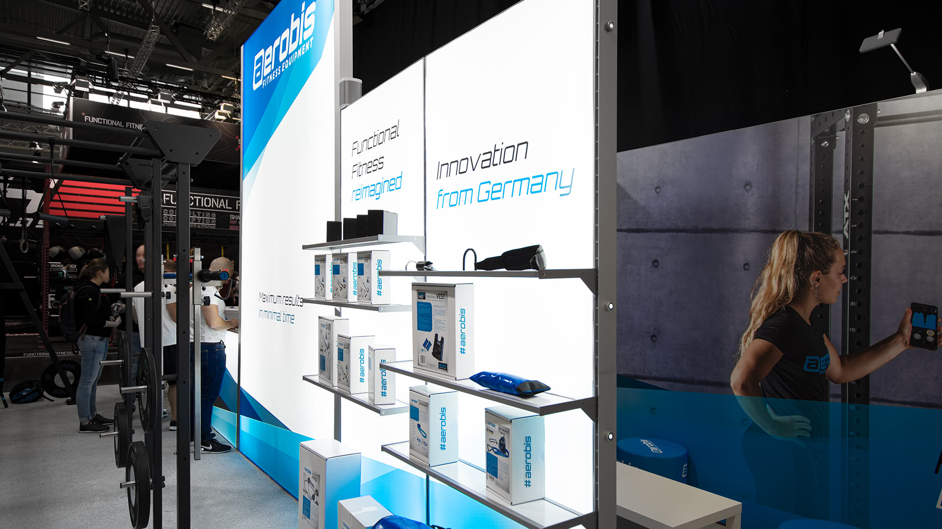 Ein Messestand für Fitnessgeräte und Zubehör mit dem Slogan Innovation from Germany. In den Regalen befinden sich blau-weiße Produkte; eine Frau benutzt ein Gerät auf der rechten Tafel. Im Hintergrund sind Besucher zu sehen, die sich unterhalten.