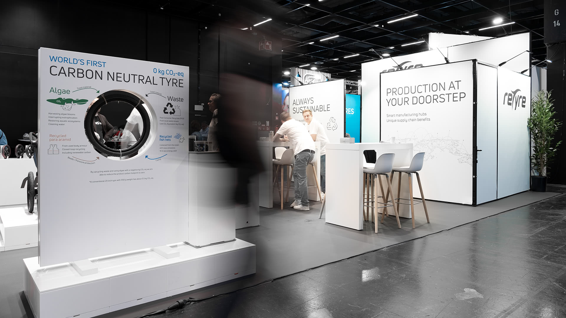 Ausstellungsstand mit dem weltweit ersten kohlenstoffneutralen Reifendisplay, mit informativen Schildern über Algen und recycelte Abfälle, mit interagierenden Menschen und modernen weißen Sitzgelegenheiten im Hintergrund.