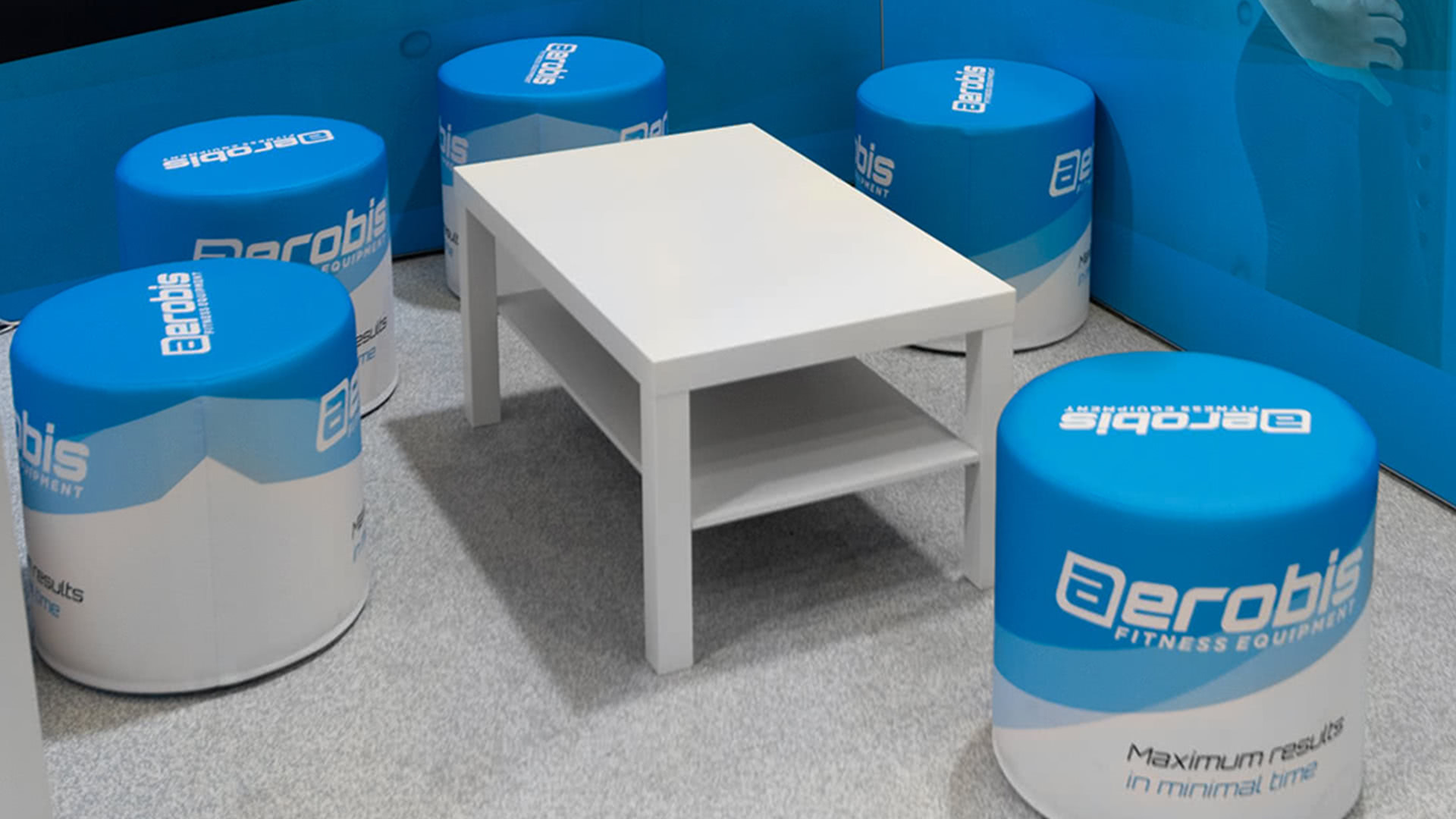 Ein weißer rechteckiger Tisch, umgeben von vier blauen und weißen runden Hockern mit dem Logo der Aerobis-Fitnessgeräte, auf einem grauen Teppich in einer Kabine oder einem Lounge-Bereich.