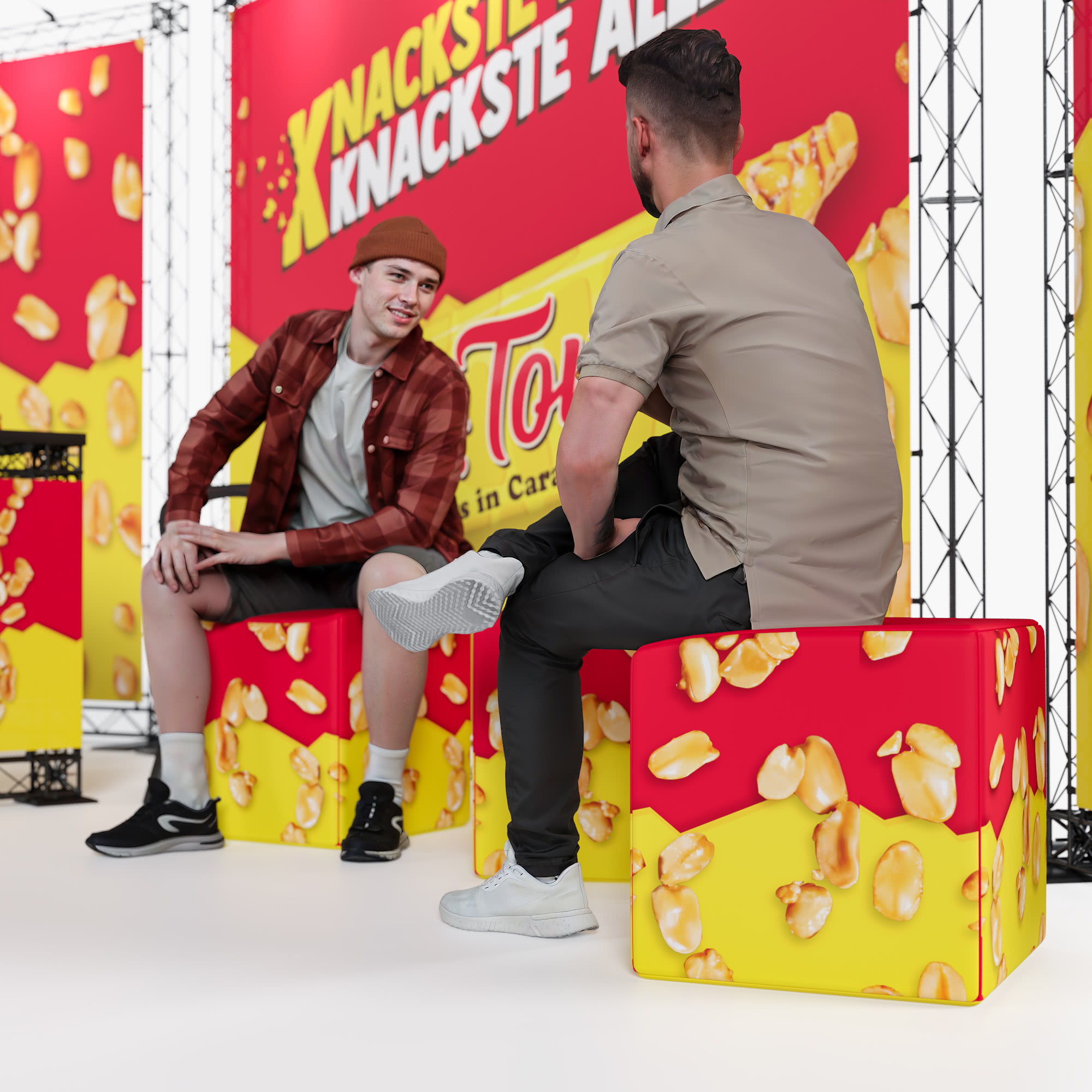 Zwei Männer sitzen auf bunten Sitzwürfeln mit Popcorn-Motiven und unterhalten sich vor einem hellen Werbehintergrund für einen Karamell-Popcorn-Snack. Der Hintergrund ist mit einem gelben und roten Branding versehen.