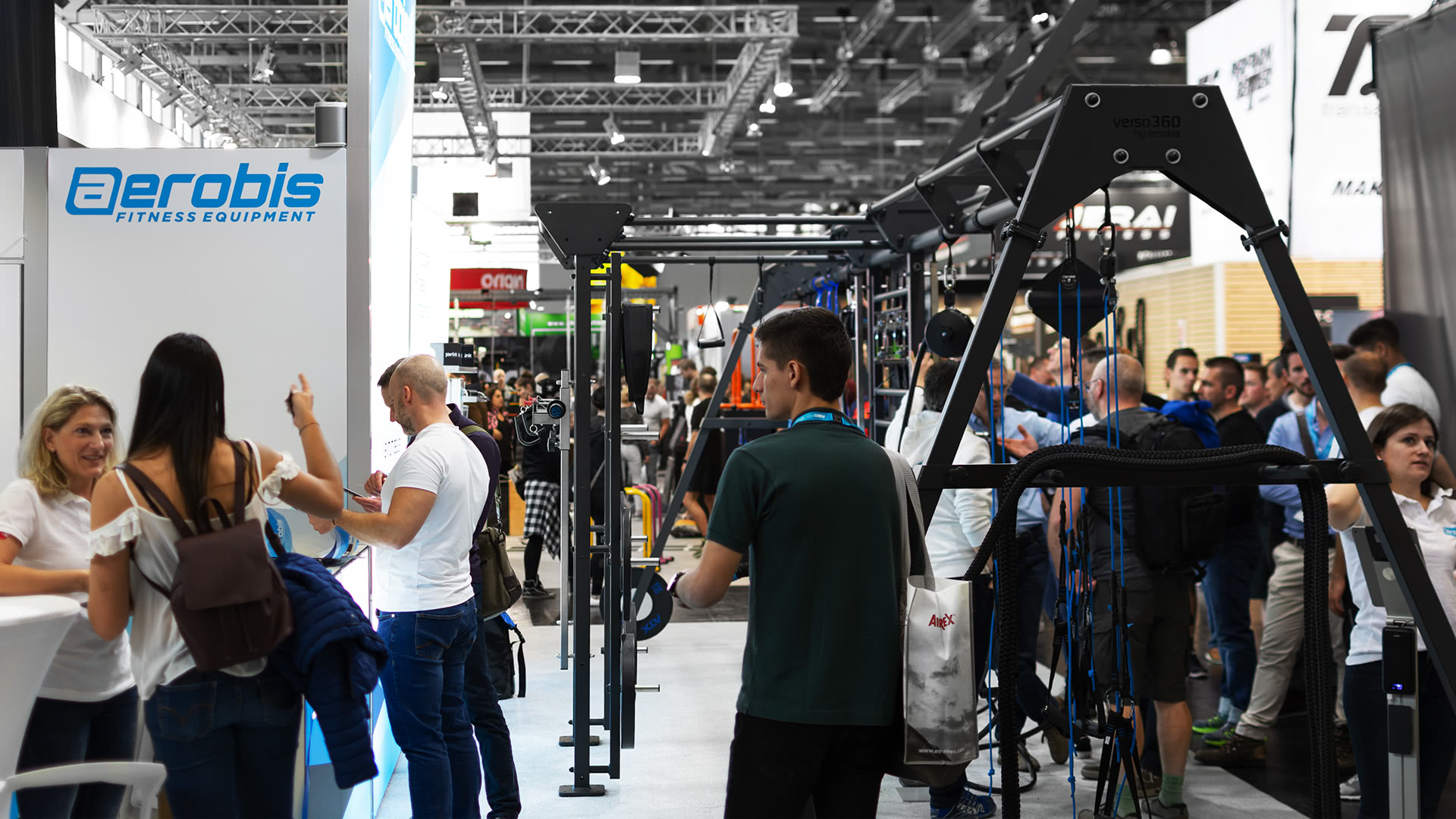Auf einer belebten Messe mit ausgestellten Fitnessgeräten und -maschinen versammeln sich Menschen und tauschen sich aus; links ist der Stand von aerobis zu sehen. Es herrscht ein reges Treiben mit zahlreichen Besuchern, die die Exponate erkunden.