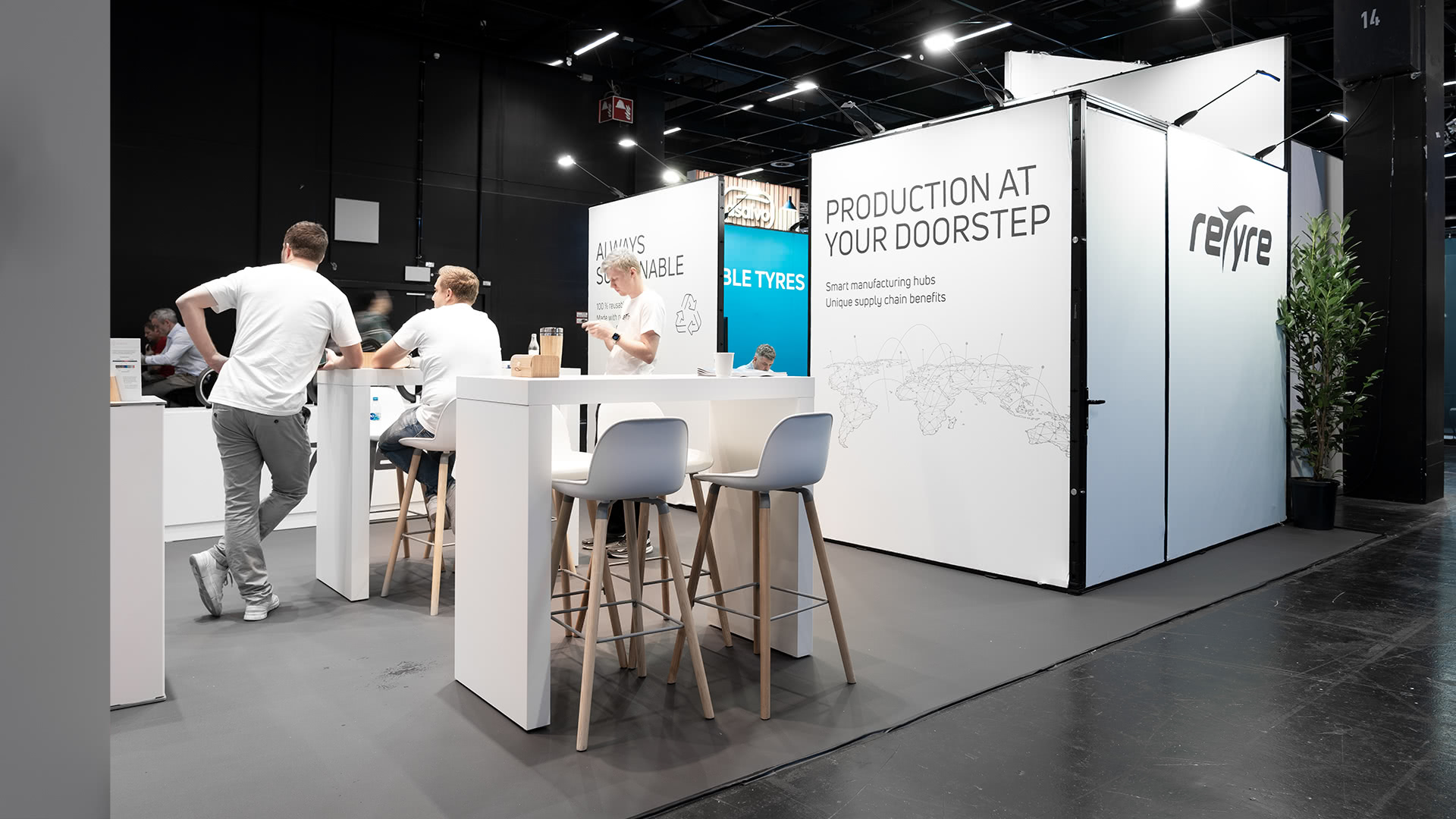 Ein moderner Messestand mit weißen Tischen und Stühlen, interagierenden Personen und einem Display mit dem Text PRODUCTION AT YOUR DOORSTEP und dem Firmennamen reTyre auf weißen Tafeln.