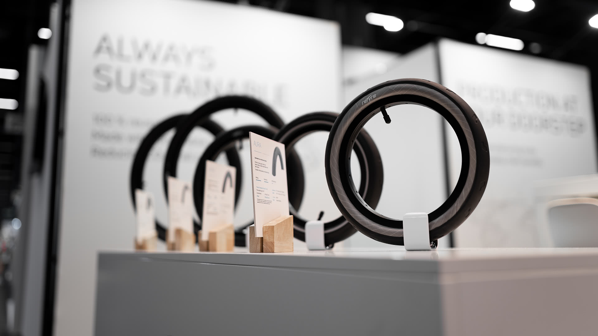 Eine Reihe aufrecht stehender Fahrradreifen auf Ständern, jeder mit einer kleinen Informationskarte davor, vor einem verschwommenen Hintergrund mit Text über Nachhaltigkeit.