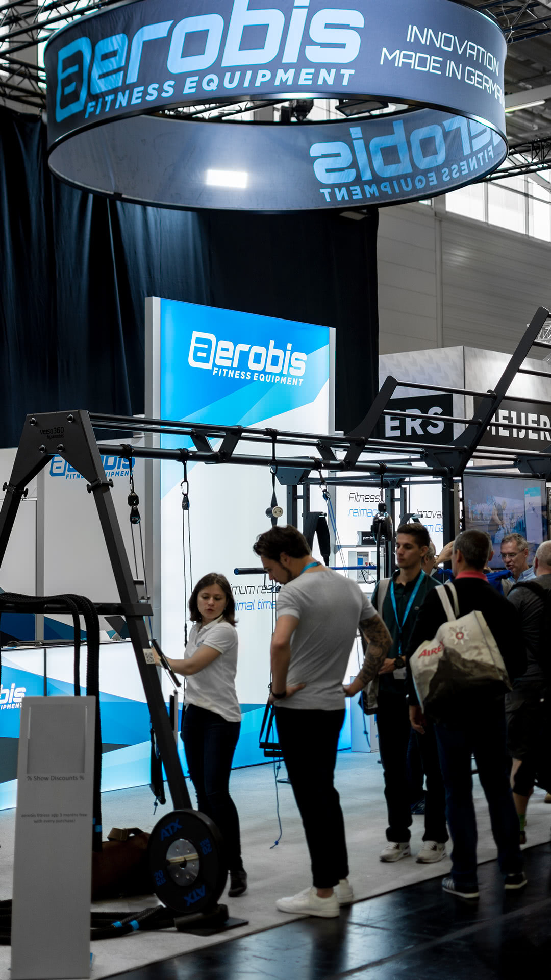 Menschen stehen an einem Ausstellungsstand für Fitnessgeräte für Aerobic, mit Displays, Markenschildern und Trainingsgeräten. Eine Frau führt Geräte vor, während andere in einer gut beleuchteten Halle zuschauen und sich austauschen.
