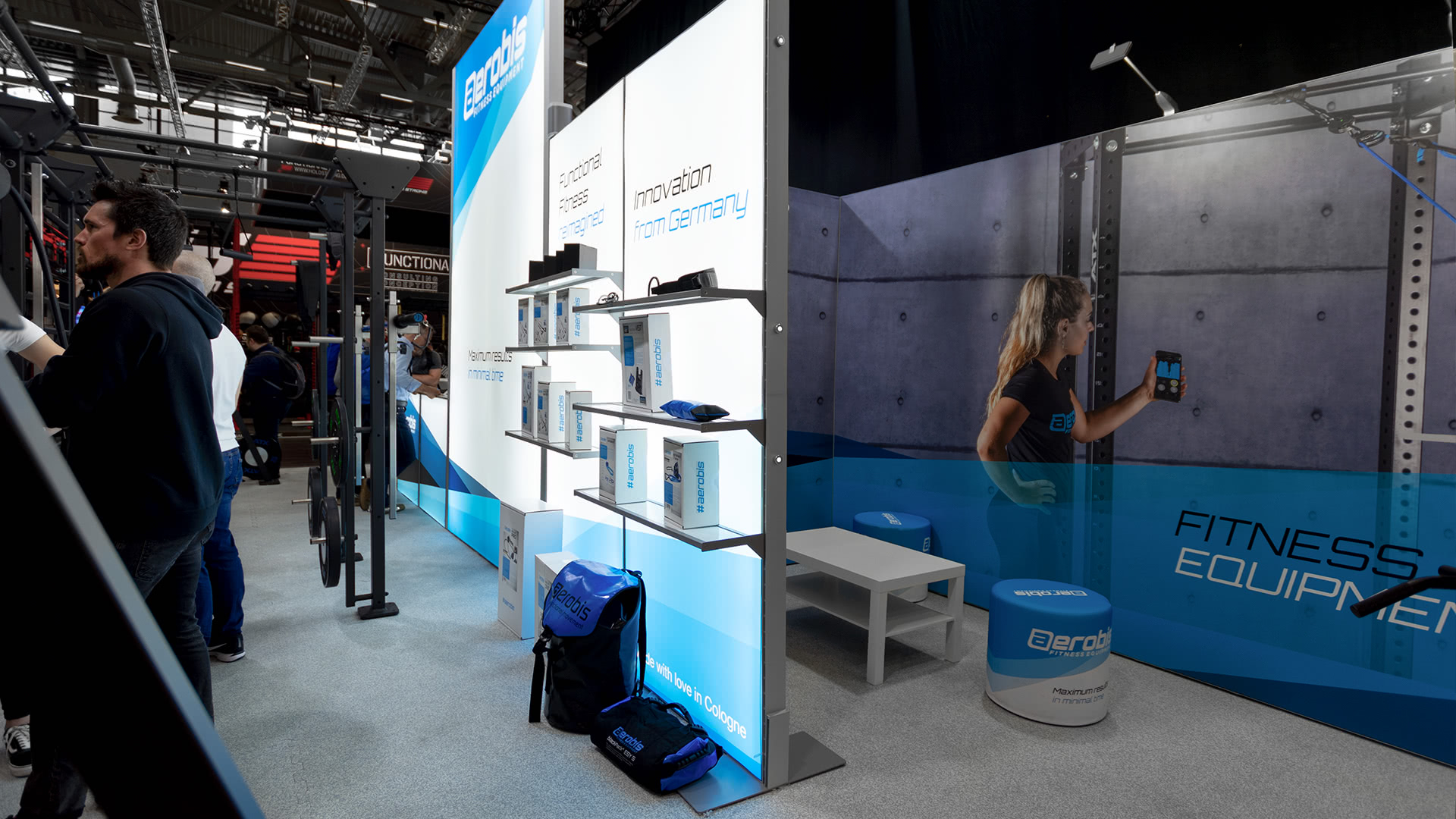 Auf einem Messestand werden Fitnessgeräte von Aerobis ausgestellt, darunter Boxen, Taschen, Mützen und Shirts. Ein großer Bildschirm zeigt eine Frau, die eine Fitness-App benutzt. In der Nähe stehen Menschen zwischen Fitnessgeräten.