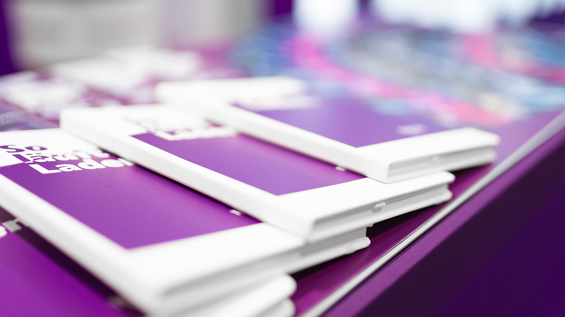 Mehrere violette und weiße Bücher oder Notizbücher sind auf einem Tisch gestapelt, wobei der unscharfe Hintergrund auf eine Veranstaltung oder Ausstellung in einem Raum hindeutet.