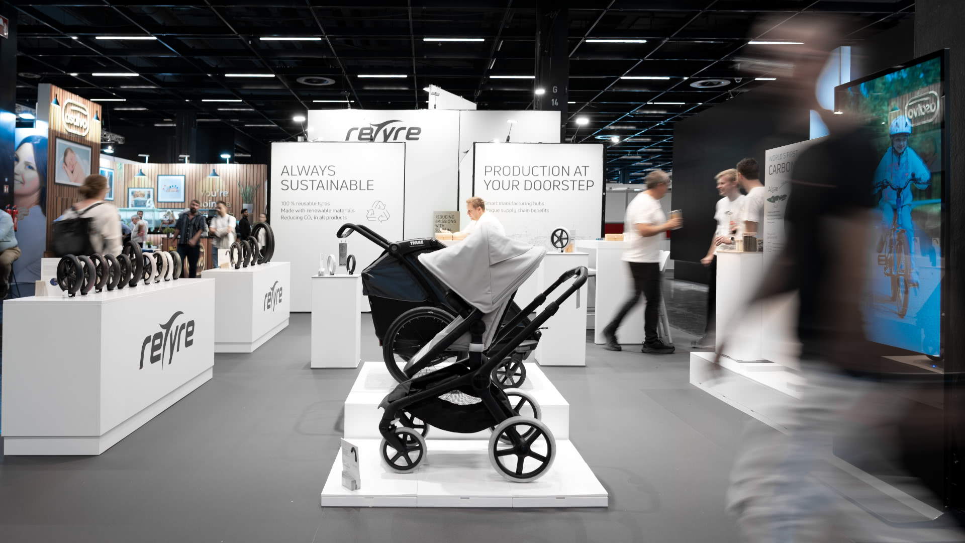 Ein Kinderwagen steht an einem Ausstellungsstand mit weißen Schildern, auf denen "Always Sustainable and Production at Your Doorstep" steht. Die Menschen gehen vorbei und betrachten andere Exponate in einem modernen, gut beleuchteten Raum.